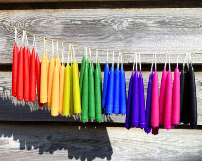 A string of colourful magical spell candles hanging outdoors against a rustic wooden wall. The colours transition in a rainbow sequence: red, orange, yellow, green, blue, purple, pink, and black.