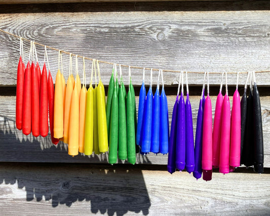 A string of colourful magical spell candles hanging outdoors against a rustic wooden wall. The colours transition in a rainbow sequence: red, orange, yellow, green, blue, purple, pink, and black.