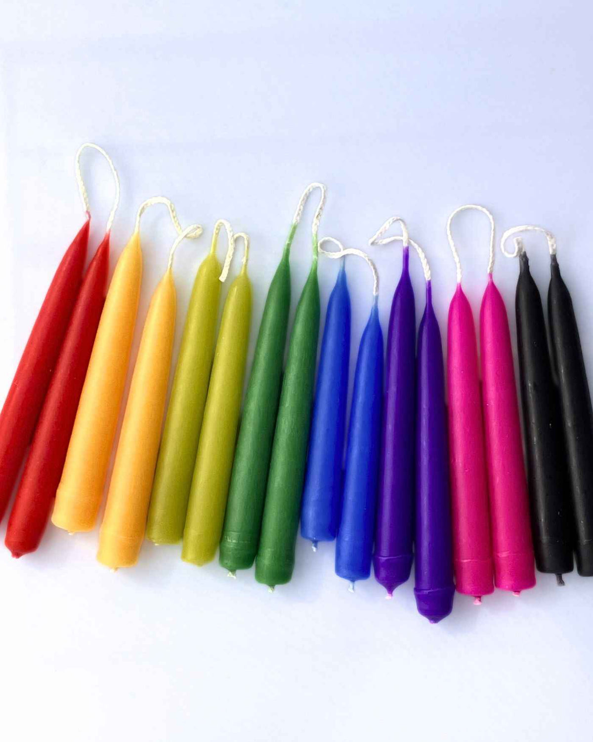 A set of colourful taper spell candles arranged in pairs in a rainbow sequence on a plain white background